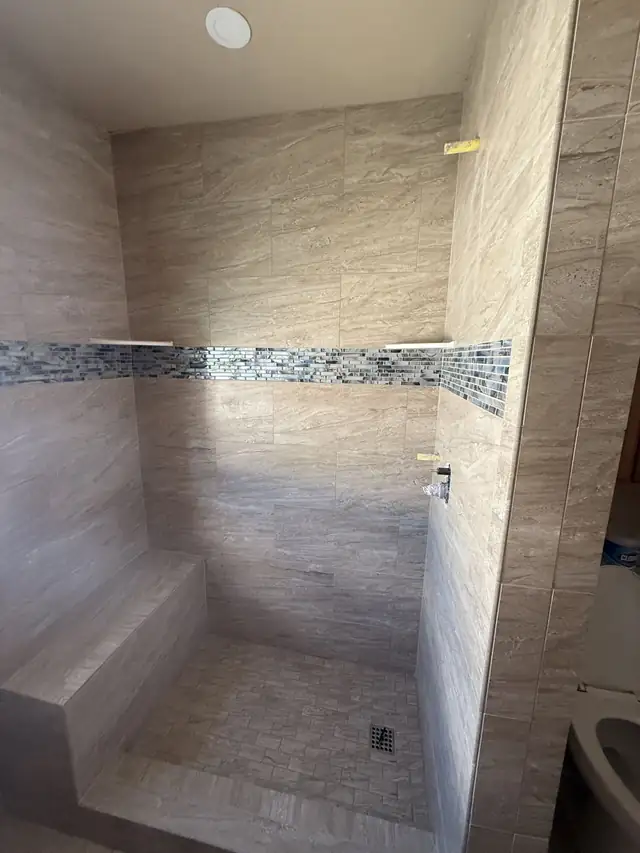 Tiled walk-in shower with large beige porcelain tile, a glass mosaic accent band, corner shelves and a built-in bench